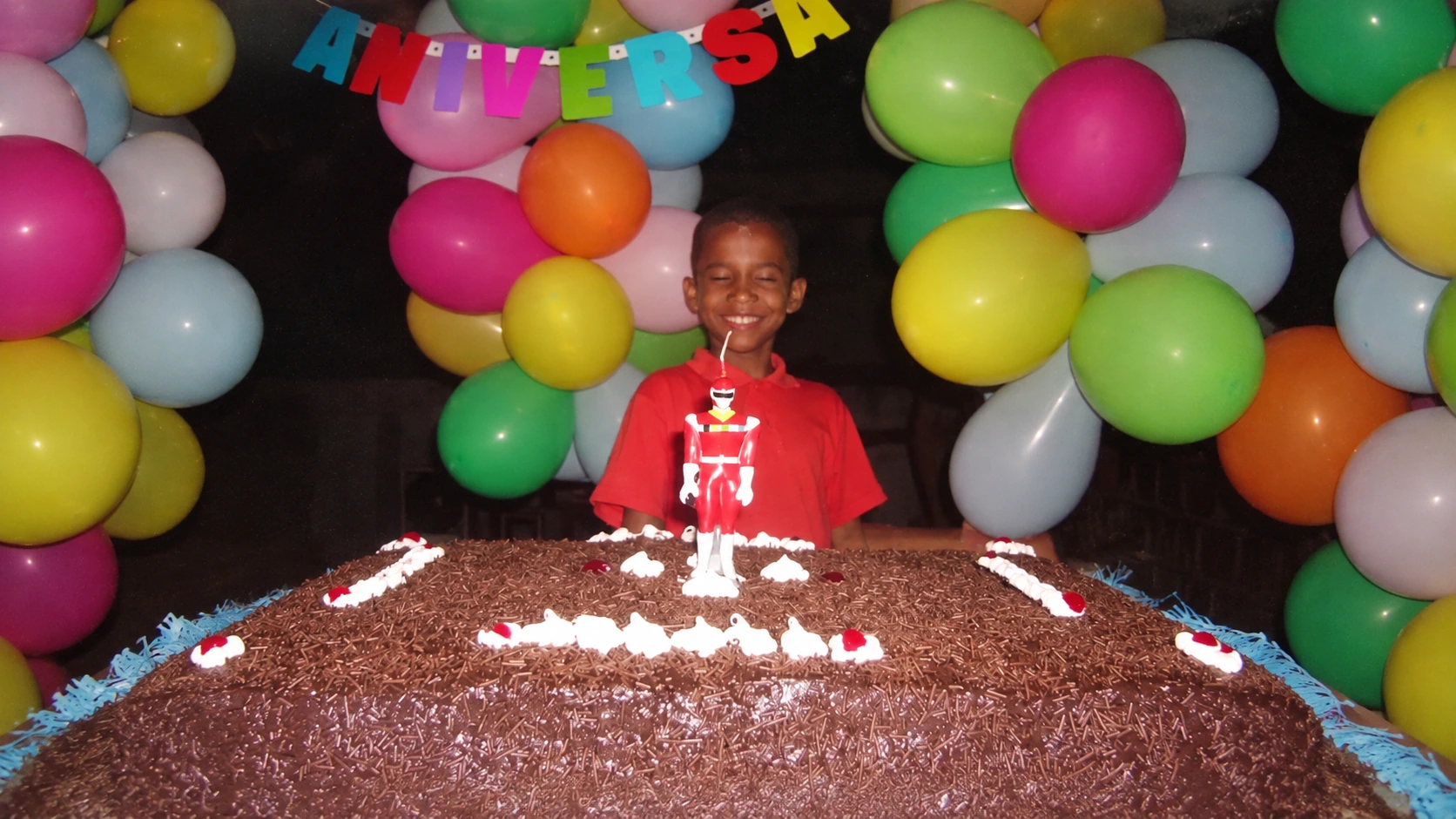DJ Matt-D ainda criança em sua festa de aniversário, diante do bolo e de balões coloridos